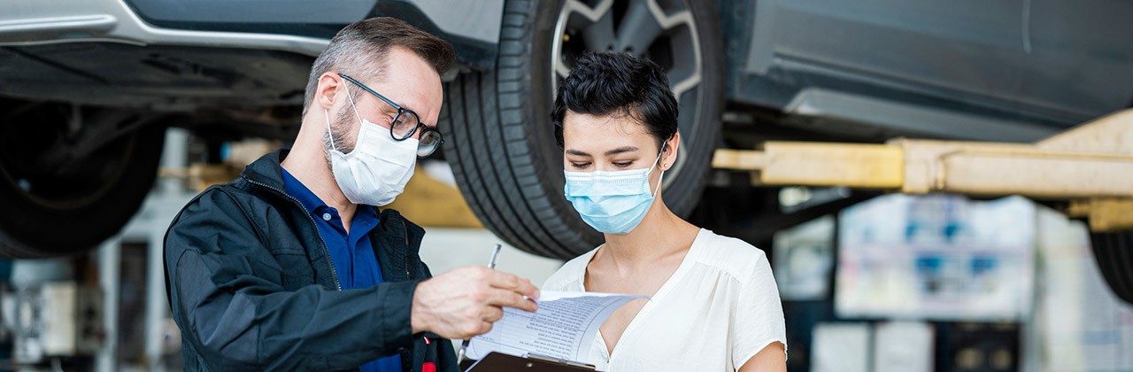 A mechanic or service rep talks to a female customer, both wearing masks, at an auto repair shop