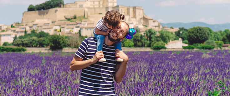 Man smiling while carrying toddler on his shoulders in a field of lavender