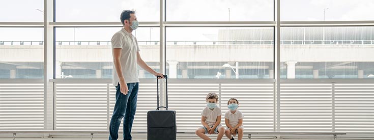 Masked man holding onto suitcase with pair of young masked boys sitting close