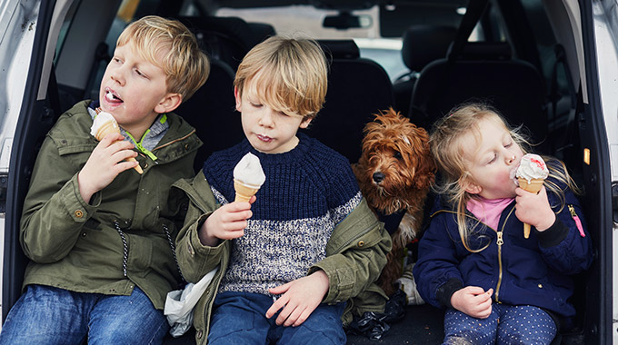 Three kids eat ice cream while sitting in the back of a car, with a dog