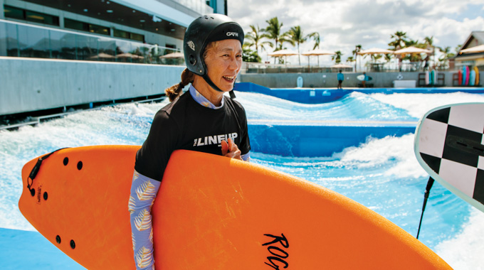 Mindy Pennybacker wearing a helmet and carrying a surf board at the artificial wave pool.