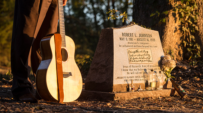 Someone holding a guitar while standing beside Robert Johnson's headstone.